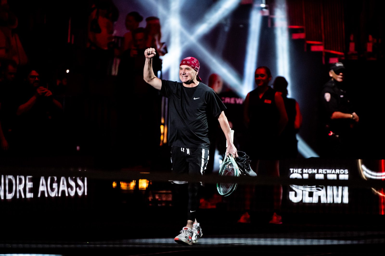 Tennis legend Andre Agassi steps out on court with lights and pyrotechnics behind him with his fist in the air and headband on.