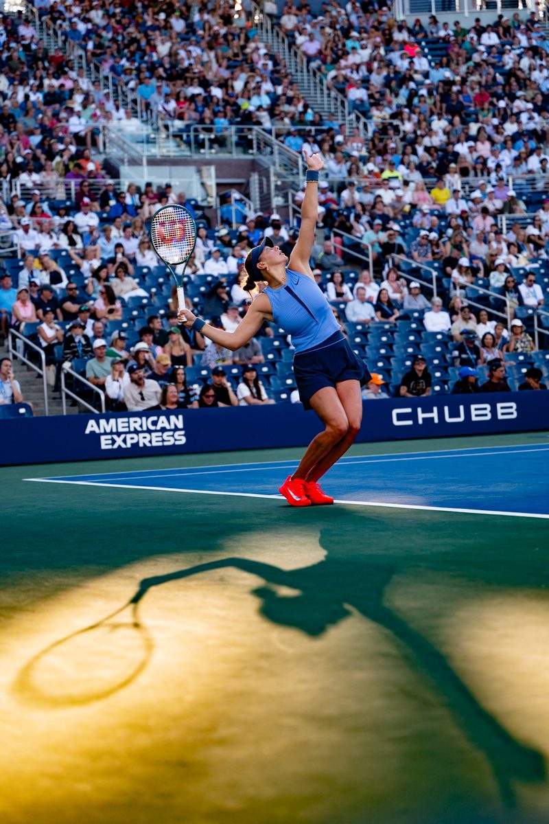 Amanda Anisimova serving at golden hour at the 2025 US Open, long shadow stretching dramatically across the court.