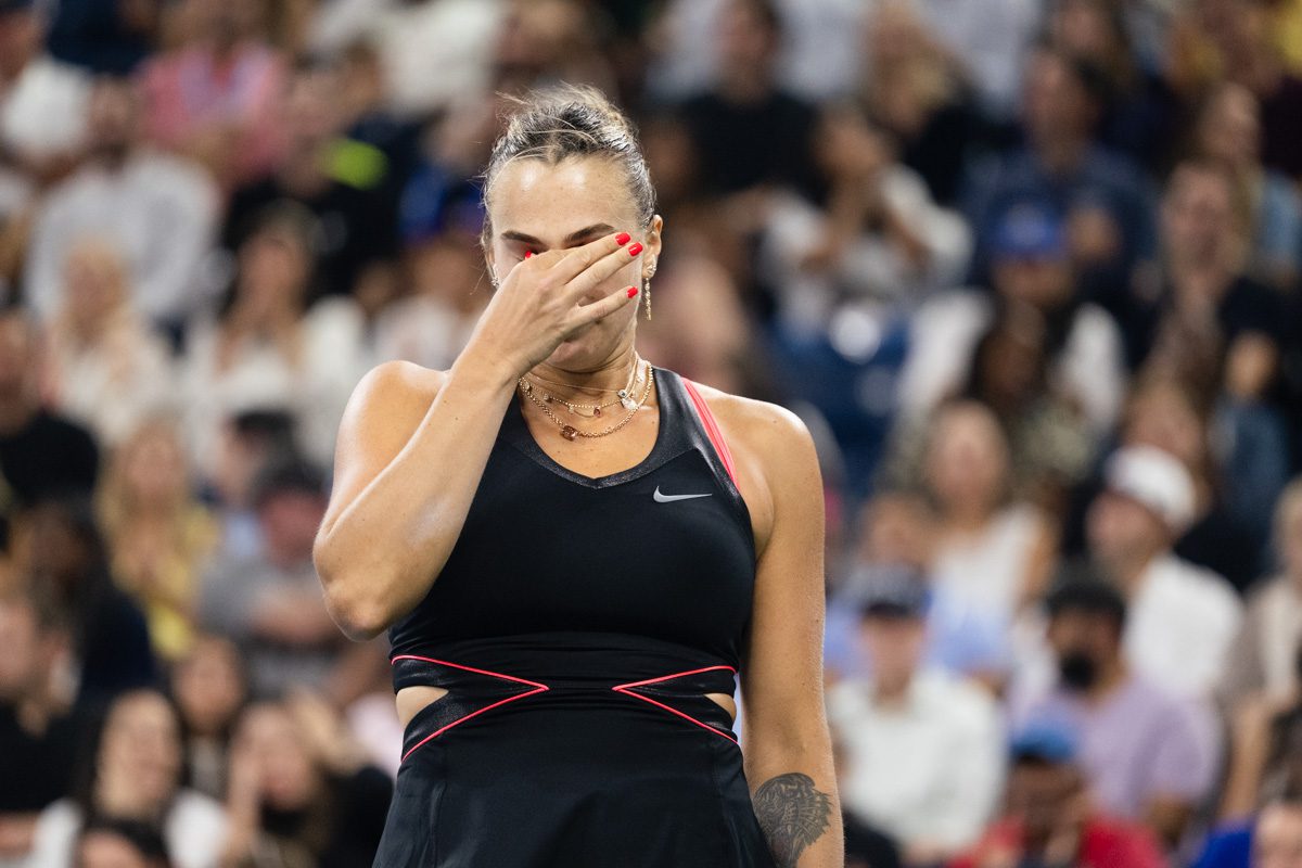 Aryna Sabalenka covers her face during an emotional moment at the 2025 US Open, crowd blurred in background.