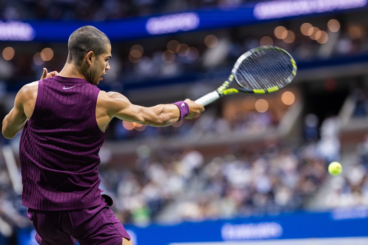 Carlos Alcaraz blasting a forehand at the 2025 US Open, muscles tense in mid-swing under stadium lights.