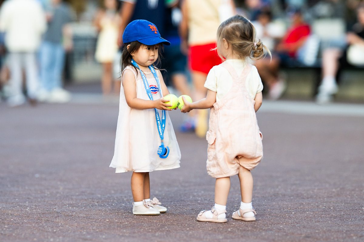 Two young girls at the 2025 US Open sharing tennis balls, fans in soft focus behind them.