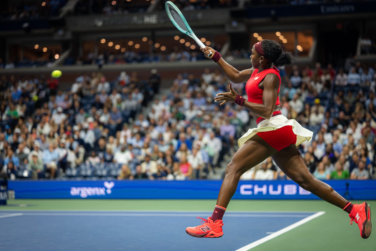Coco Gauff airborne hitting a forehand in bright red kit during a 2025 US Open night match.