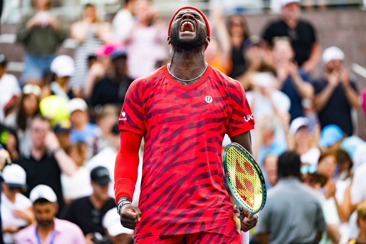 Frances Tiafoe roars in celebration on Grandstand court at the 2025 US Open, arms flexed in bright red kit as fans cheer.