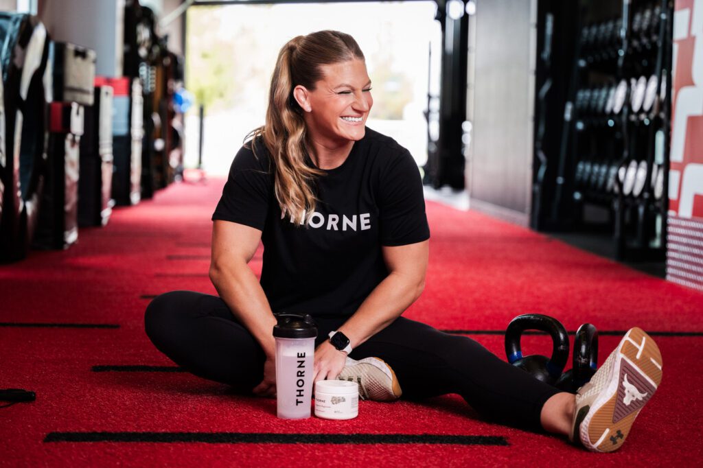 Kayla Harrison seated on the red floor at the UFC Performance Institute with Thorne shaker and weights beside her during a campaign photo shoot.