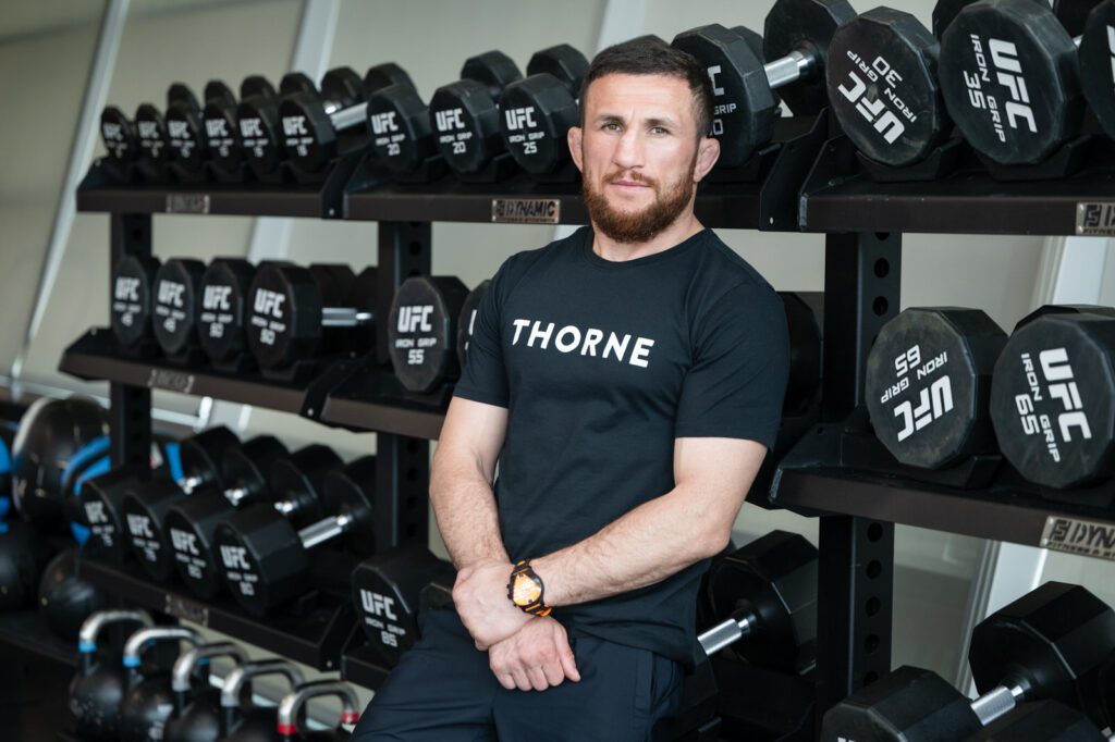 Merab Dvalishvili posing beside UFC dumbbells during a Thorne supplements campaign at the UFC Performance Institute in Las Vegas.