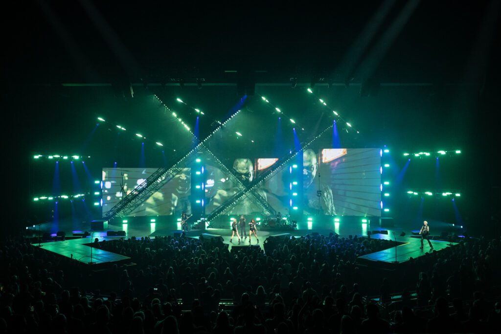 Wide frame showing Mötley Crüe surrounded by green and blue light at Dolby Live, with diagonal trusses and LED walls emphasizing the set’s geometry.