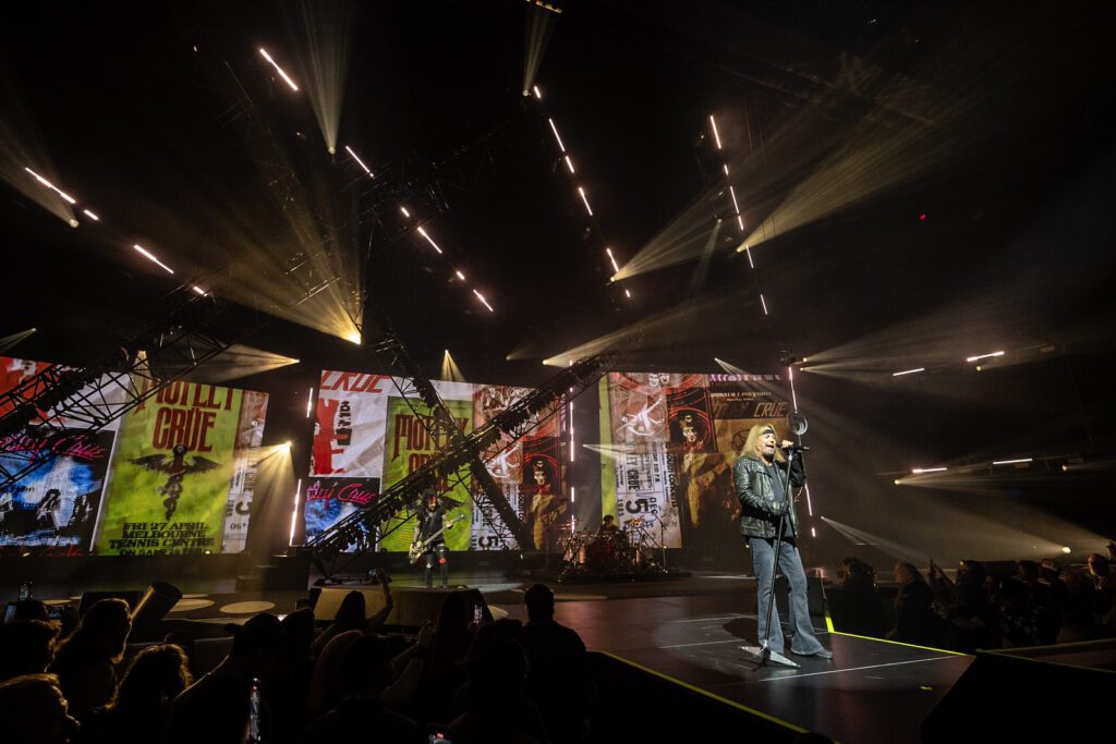 A wide view of Mötley Crüe performing in front of vintage-style poster graphics and angled trusses, captured in warm light and haze during their Park MGM residency.