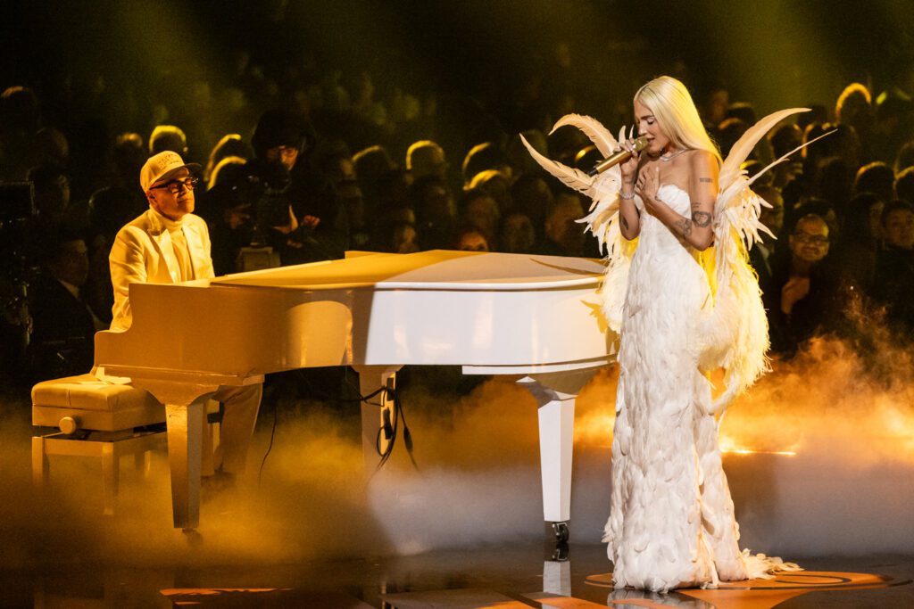A performer sings beside a white grand piano while wearing a white feathered gown during a Latin Grammy stage performance.