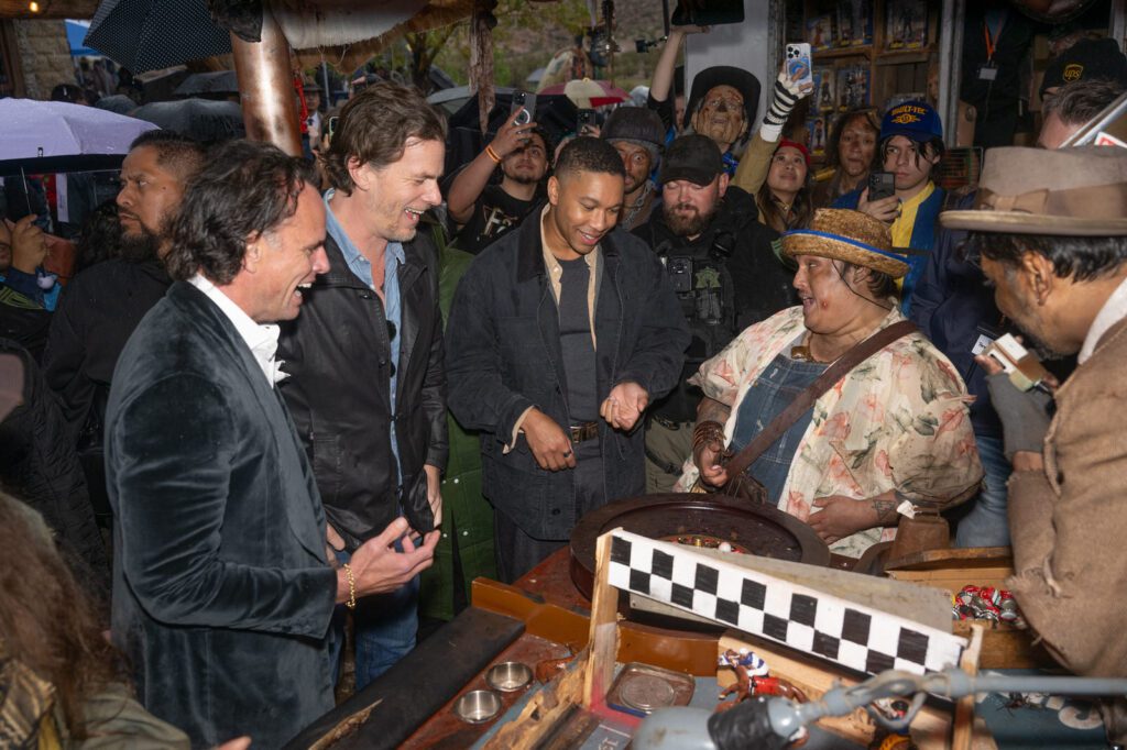 A group gathers around a tabletop game as three men watch and smile during the Fallout-themed event.
