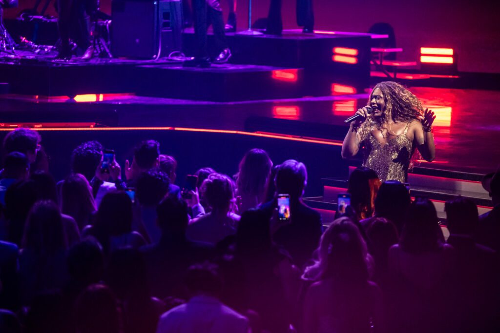 Liniker A performer sings during the Latin Grammy telecast, illuminated by purple lighting with the audience visible below.
