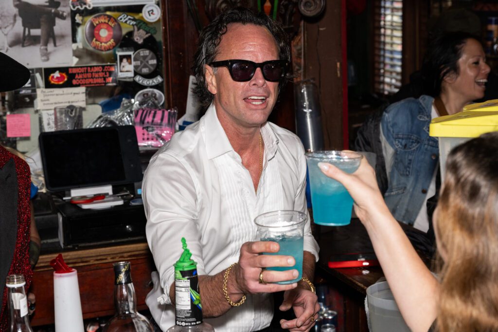 A man in sunglasses holds a blue drink while raising his glass toward a fan inside the packed Pioneer Saloon bar.