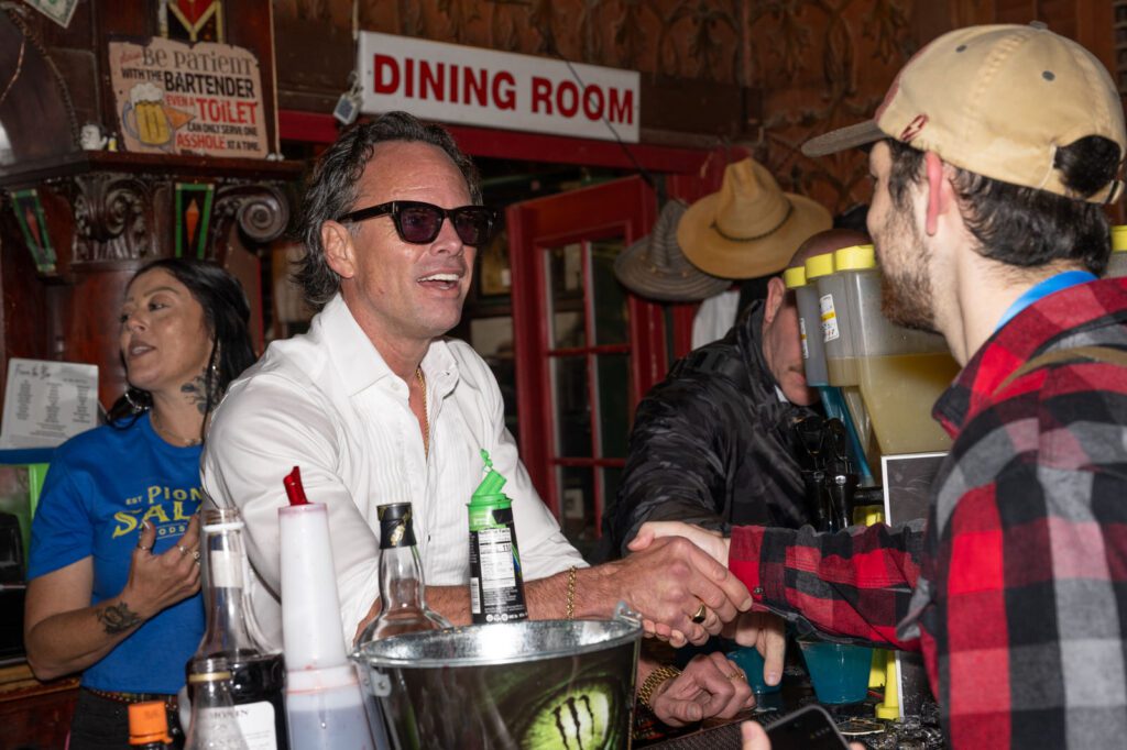 A man in sunglasses stands behind the Pioneer Saloon bar shaking hands with an attendee during the Fallout celebration.
