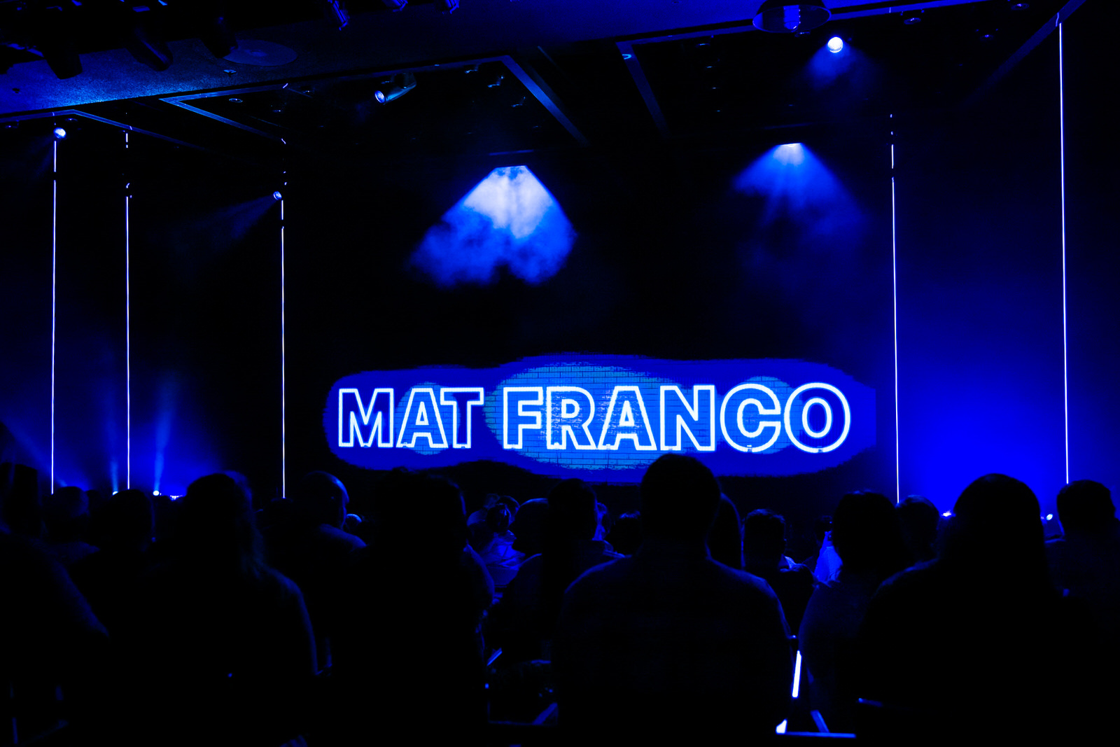Silhouetted audience members take their seats inside the intimate Mat Franco Theater at the LINQ Hotel in Las Vegas, bathed in deep blue light, with a glowing neon Mat Franco sign illuminating the stage screen ahead of curtain