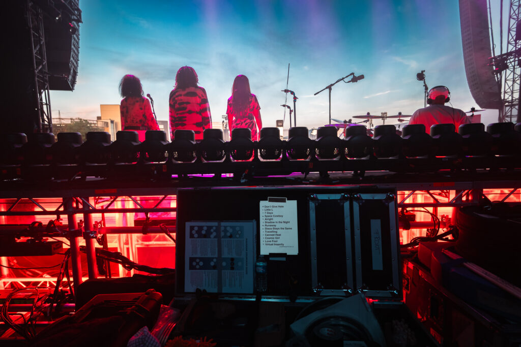 Three backing vocalists stand silhouetted against a sunset sky on stage during Jamiroquai's Heels of Steel Tour show in Luxembourg, a printed setlist visible on the equipment case in the foreground.