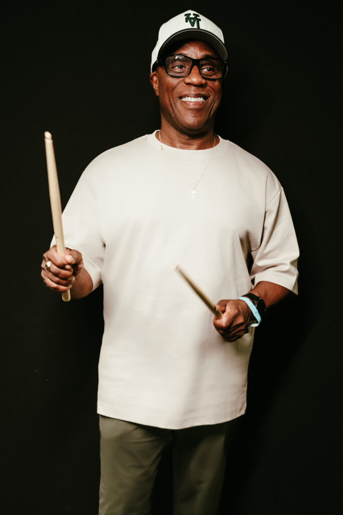 Derrick McKenzie, drummer for Jamiroquai, poses backstage holding drumsticks and smiling, wearing a white cap and glasses, during a quick portrait session before the Heels of Steel Tour show in Luxembourg.
