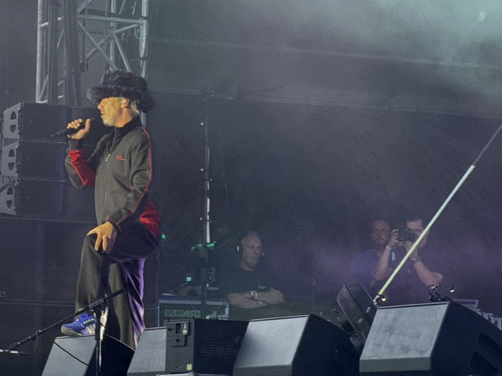 Jamiroquai frontman Jay Kay sings into the microphone wearing his signature furry hat, photographed from the side under purple stage lighting during the Heels of Steel Tour stop in Luxembourg.